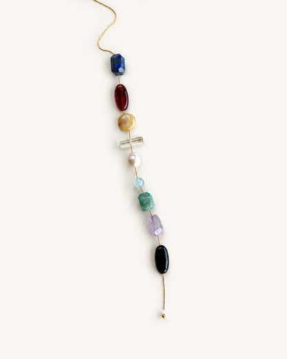 A delicate gold beaded necklace featuring a vertical line of colorful gemstone beads in blue, red, gold, white, light blue, green, purple, and black, displayed on a solid off-white background.