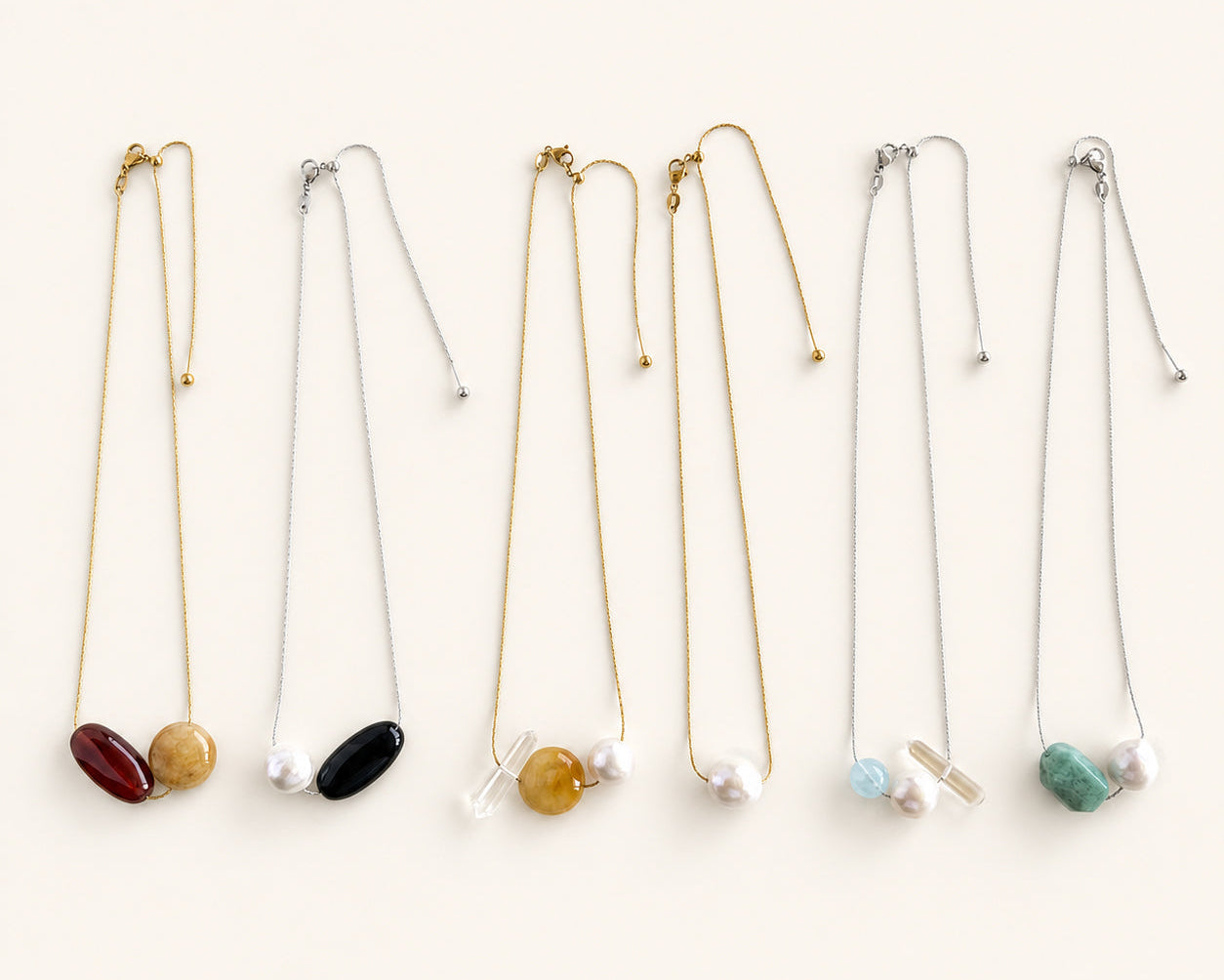 Six delicate necklace chains arranged in a row on a light cream background, each featuring a small oval stone pendant in different colors including red, yellow, black, gold, light blue, and green.