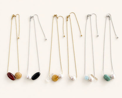 Six delicate necklace chains arranged in a row on a light cream background, each featuring a small oval stone pendant in different colors including red, yellow, black, gold, light blue, and green.