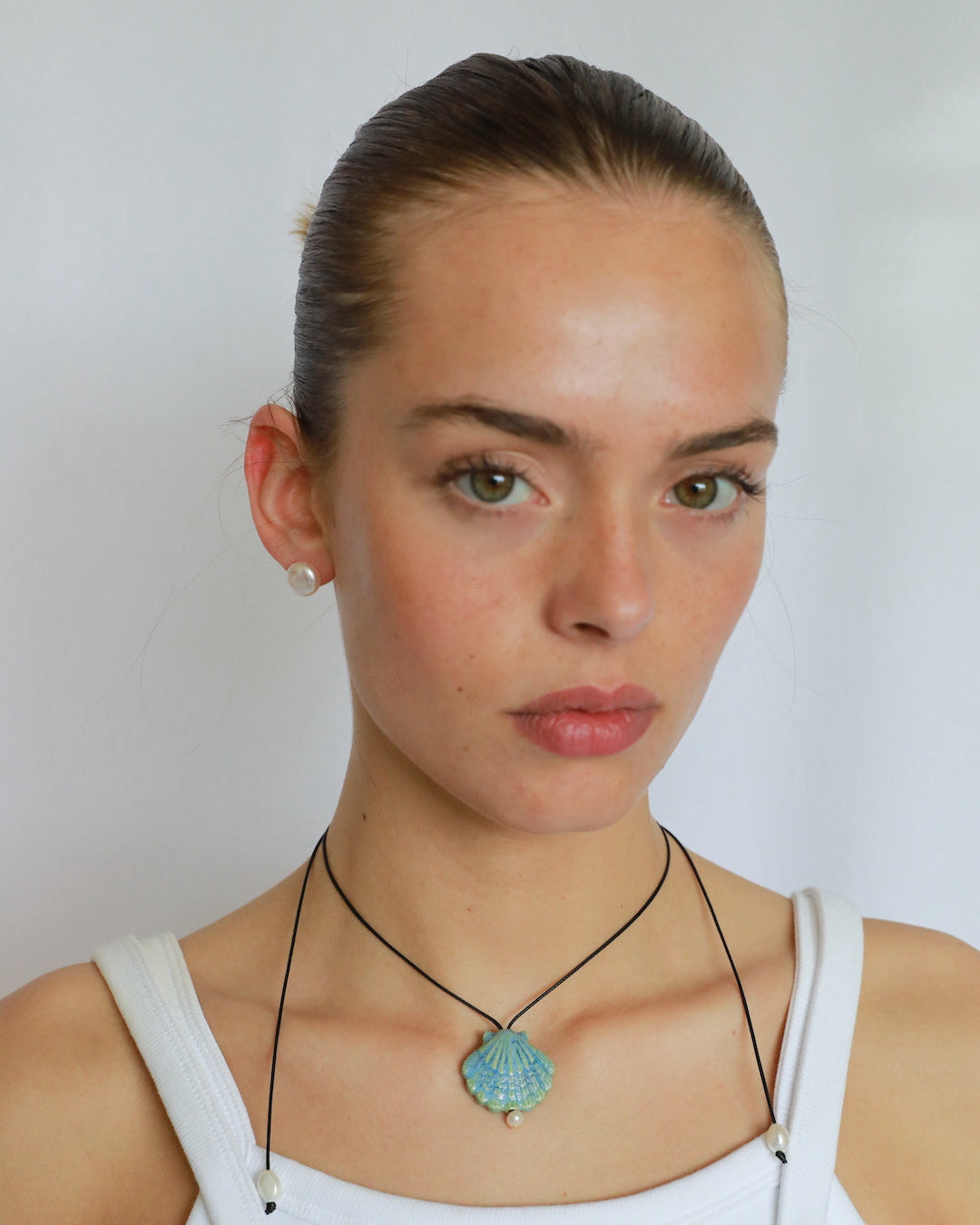 A woman with her hair pulled back wears a white tank top and a black cord necklace with a round turquoise-blue pendant, photographed against a plain light gray background.