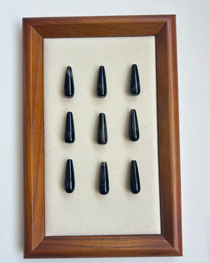 A framed display of nine glossy black teardrop-shaped stones arranged in a 3x3 grid on a beige backing, set inside a medium brown wooden frame against a plain light background.