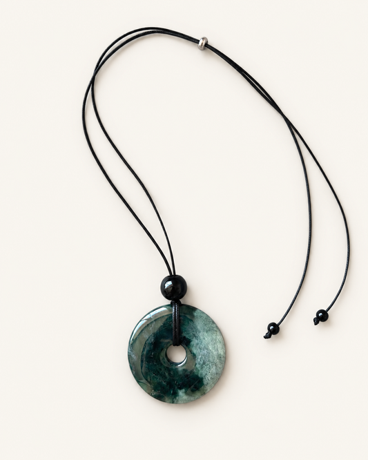 A circular green jade pendant necklace with a black cord and adjustable slider, displayed on a light cream background.