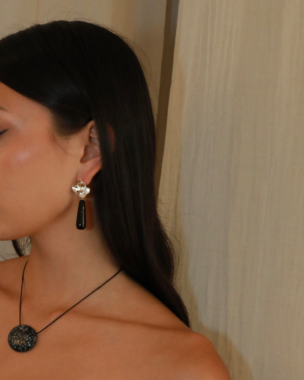 A close-up side profile of a woman with long dark hair wearing a black teardrop pendant earring and a black circular pendant necklace against a beige curtain background.