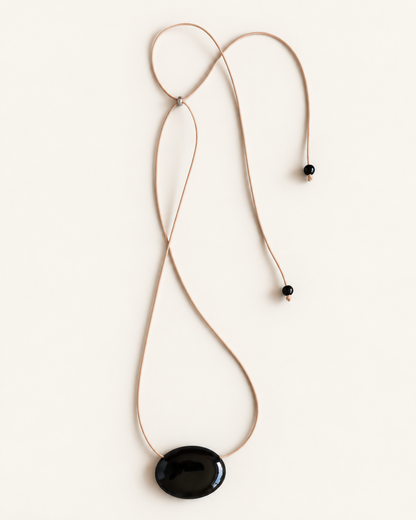 A minimalist necklace featuring a large, smooth black oval pendant suspended from a thin tan cord with small black beads, laid flat on a light cream background.