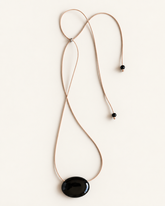 A minimalist necklace featuring a large, smooth black oval pendant suspended from a thin tan cord with small black beads, laid flat on a light cream background.