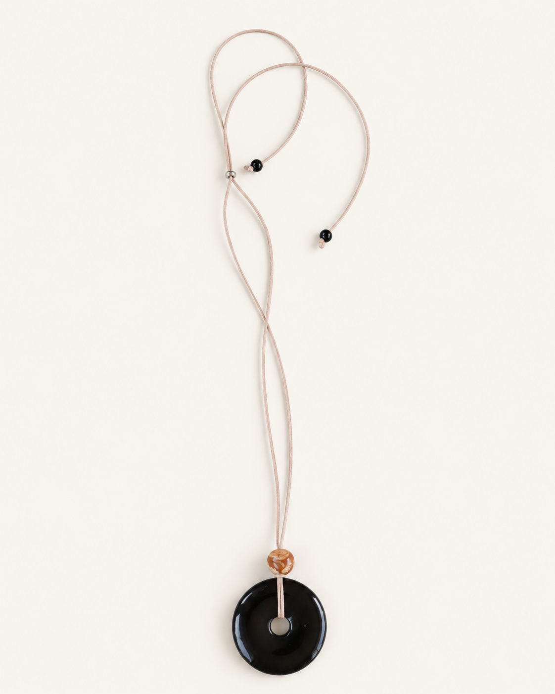 A minimalist necklace featuring a large, smooth black circular pendant on a thin beige cord, accented by a small tan bead and two tiny black beads, shown against a solid off-white background.