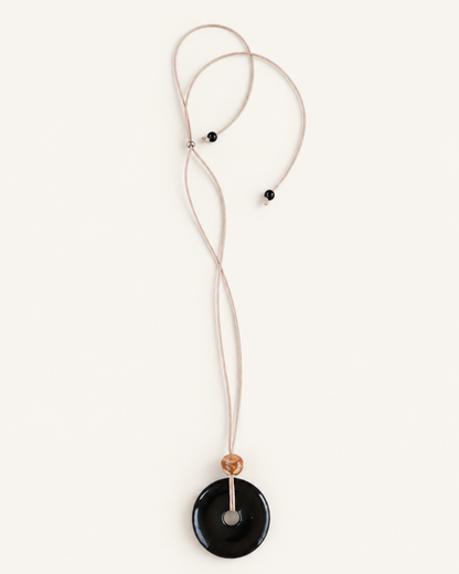 A minimalist necklace featuring a large, smooth black circular pendant on a thin beige cord, accented by a small tan bead and two tiny black beads, shown against a solid off-white background.
