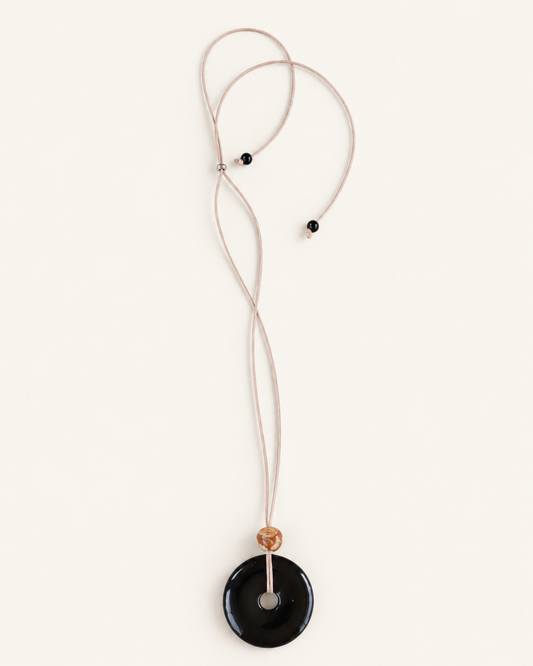 A minimalist necklace featuring a large, smooth black circular pendant on a thin beige cord, accented by a small tan bead and two tiny black beads, shown against a solid off-white background.