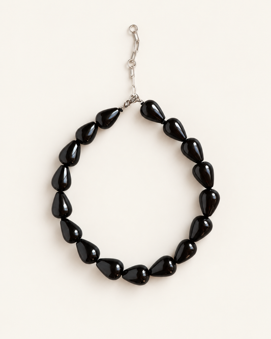 A beaded necklace made of glossy black teardrop-shaped beads with a silver-tone clasp, displayed on a light cream background.