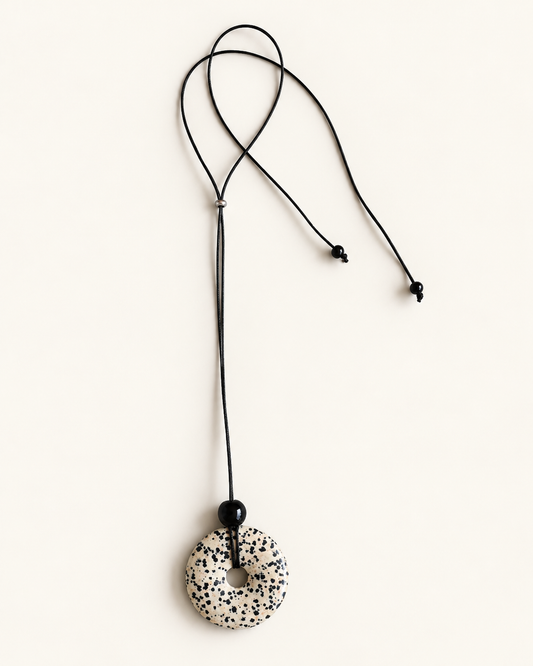 A black adjustable cord necklace featuring a circular donut-shaped pendant with a speckled black and white pattern, displayed on a clean off-white background.