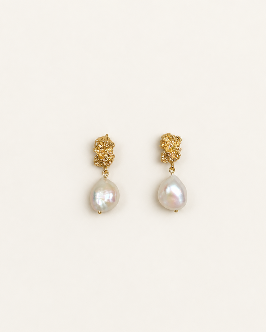 A pair of gold dangle earrings featuring textured gold studs and teardrop-shaped white baroque pearls, displayed on a clean off-white background.