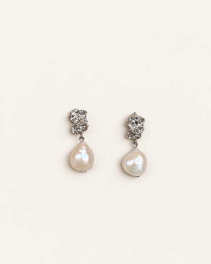 A pair of silver-toned drop earrings featuring a textured hoop at the top and a teardrop-shaped white baroque pearl hanging below, set against a plain off-white background.