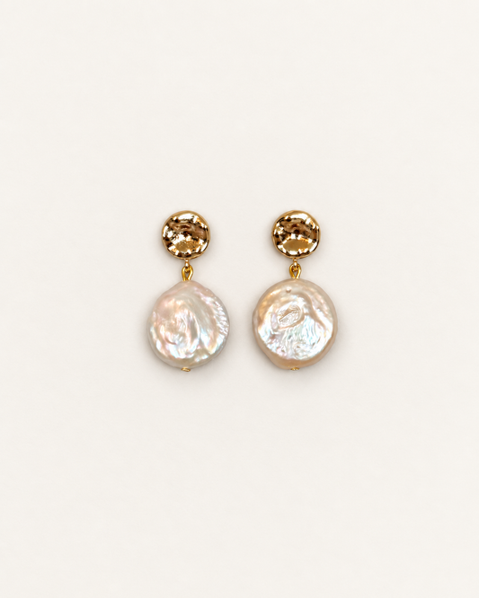 A pair of gold drop earrings featuring a faceted round gold top and a hanging irregular baroque pearl, shown on a plain off-white background.
