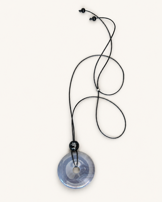 A circular translucent blue pendant necklace with a black cord and black beads, displayed on a clean off-white background.