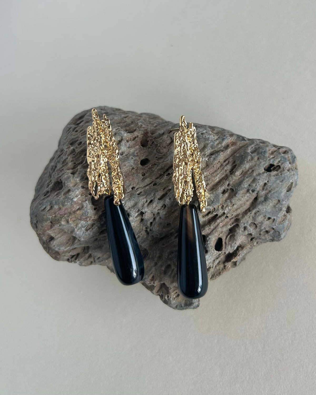 A pair of statement earrings with textured gold leaf-like tops and glossy black teardrop pendants, displayed on a rough gray rock against a light beige background.