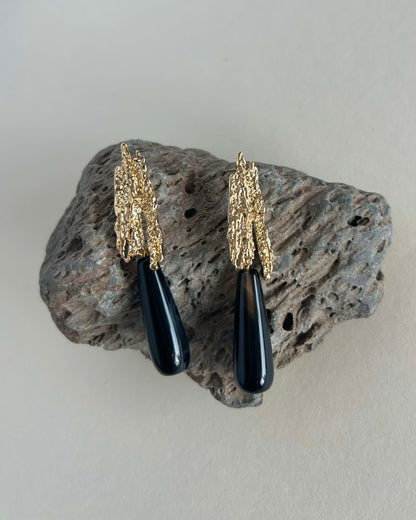 A pair of statement earrings with textured gold leaf-like tops and glossy black teardrop pendants, displayed on a rough gray rock against a light beige background.