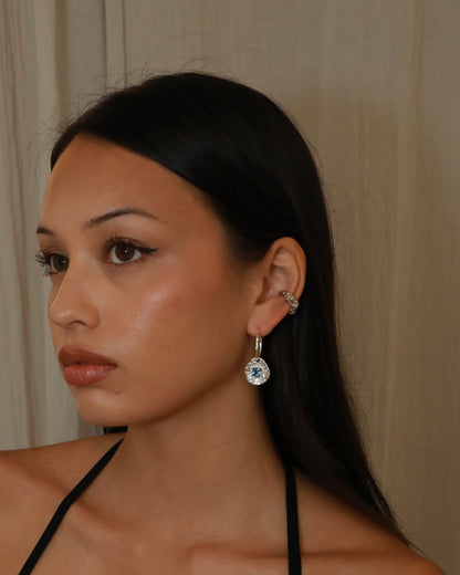 A close-up of a woman with long dark hair wearing a silver-toned dangle earring with a clear faceted gemstone. The background is a soft beige curtain.