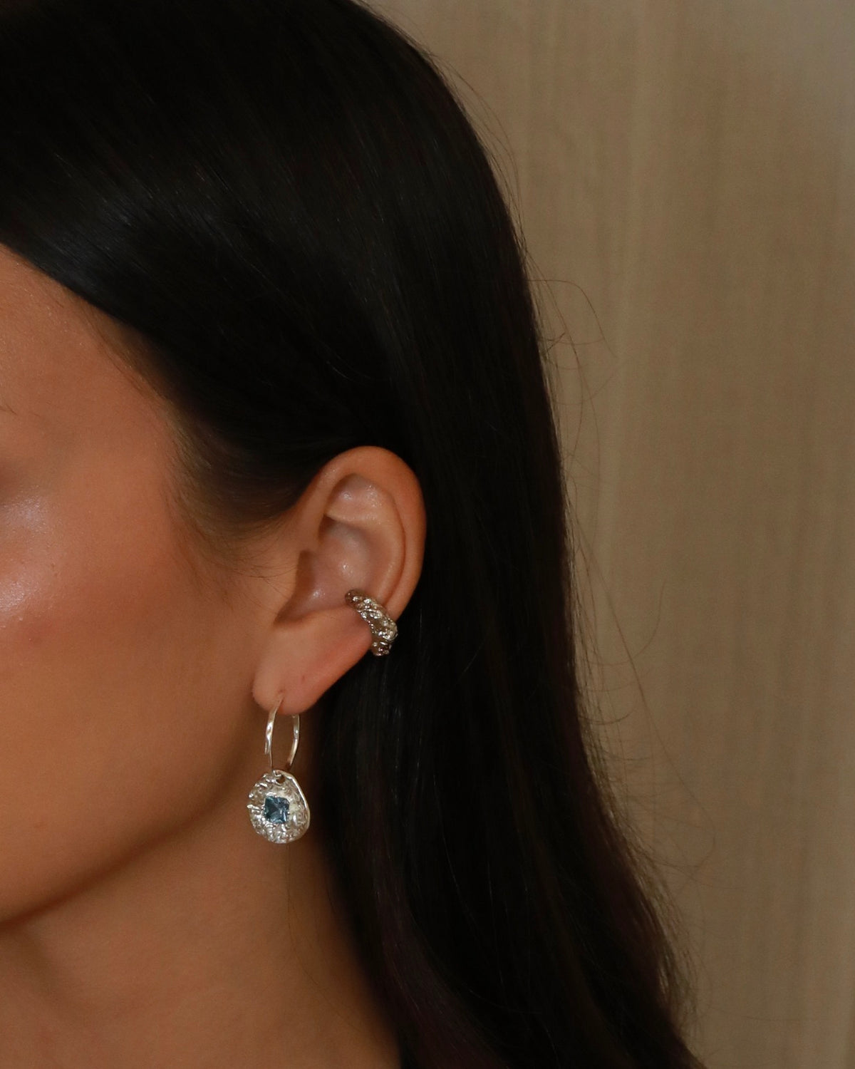 A close-up of a model's ear wearing a silver-toned hoop earring with a round blue gemstone surrounded by small clear stones, along with a silver ear cuff.