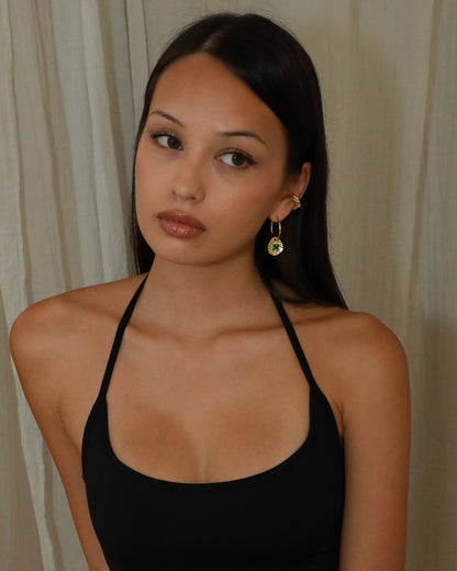 A woman with long dark hair wearing a black halter top and gold-toned dangle earrings, photographed against a light beige curtain background.