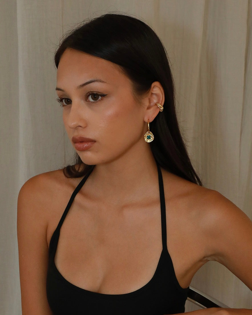A woman with long dark hair wearing a black halter top and a gold hoop earring with a green gemstone and textured earcuff, photographed against a light beige curtain background.