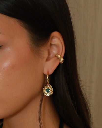 A close-up of a model's ear wearing a gold huggie hoop earring with a dangling teardrop charm featuring a green gemstone surrounded by small clear crystals and textured earcuff