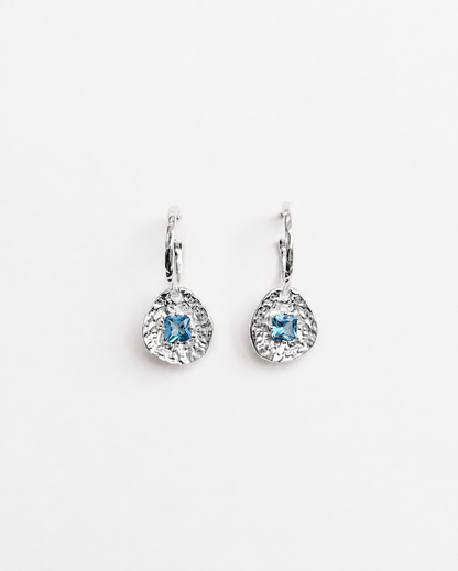 Cleo silver-plated brass coin drop earrings with hammered texture and square blue tourmaline CZ stones on a soft white background.